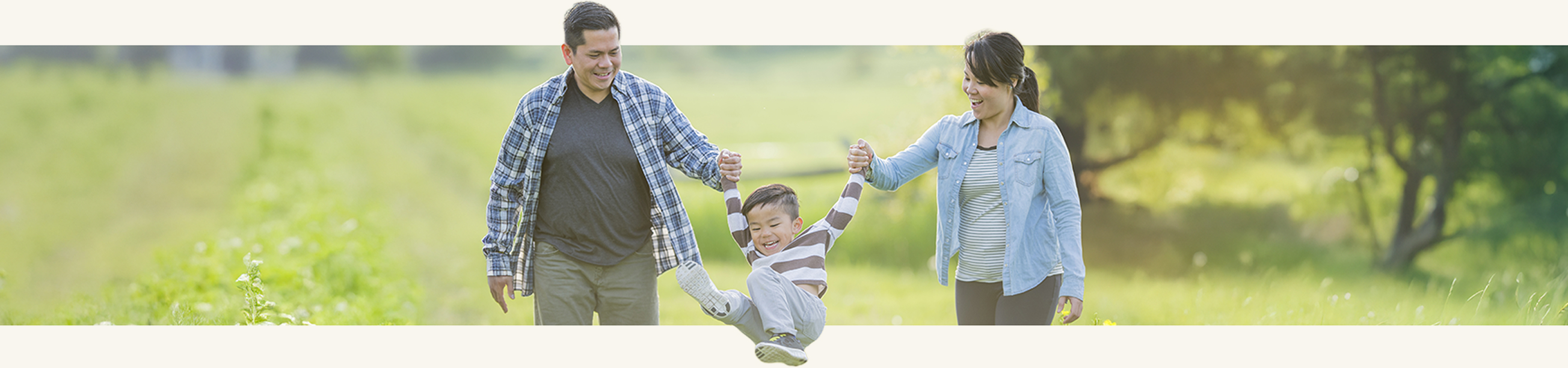 Smiling parents swinging their laughing young child by the arms while walking together through a grassy field on a sunny day.